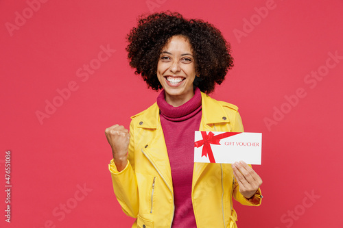 Charming vivid excited young curly black latin woman 20s wears yellow jacket hold gift certificate coupon voucher card for store doing winner gesture isolated on plain red background studio portrait.