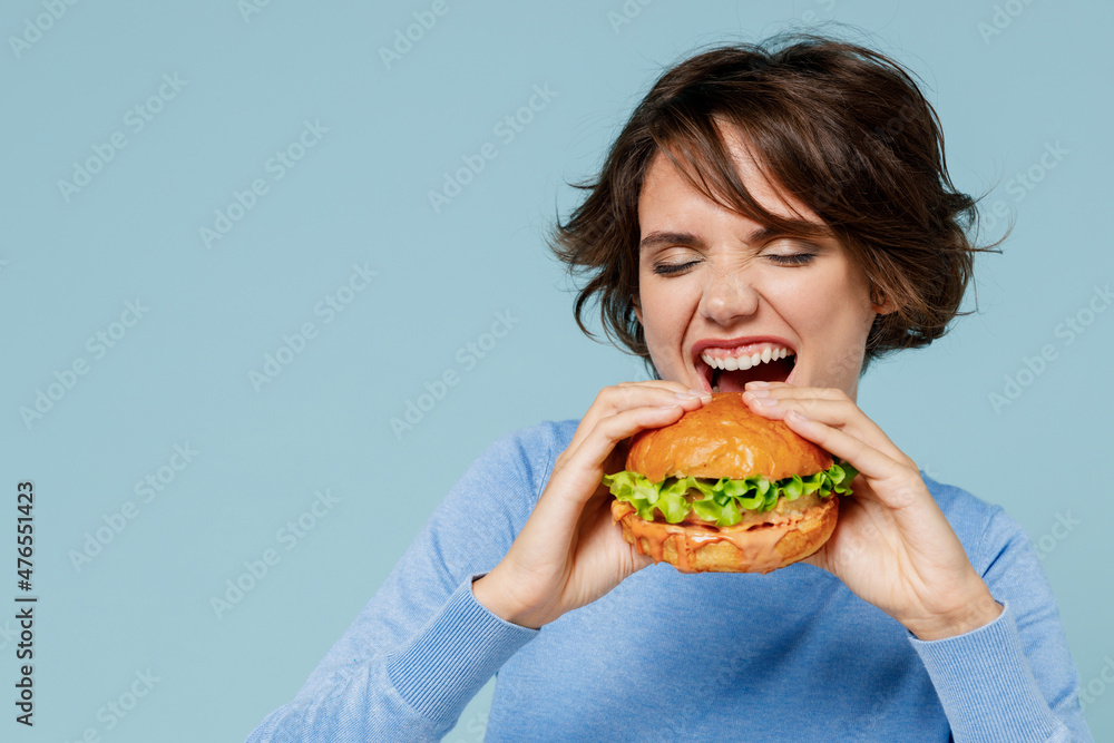 Young hungry caucasian fun cool woman 20s wearing casual sweater biting eating burger isolated on plain pastel light blue color background studio portrait People lifestyle unhealthy junk food concept