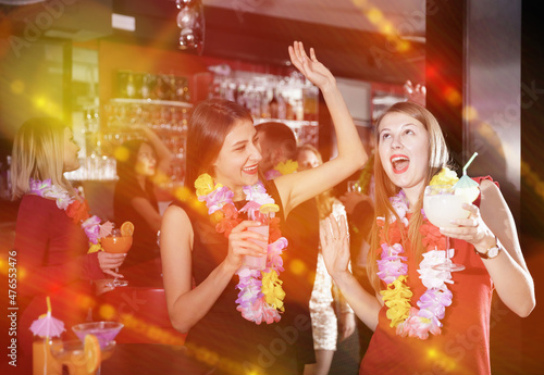 Cheerful girls dancing and toasting drinks on Hawaiian party in bar