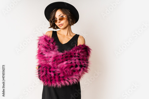 Graceful skinny woman in stylish winter fluffy pink  coat and black hat posing on white background.