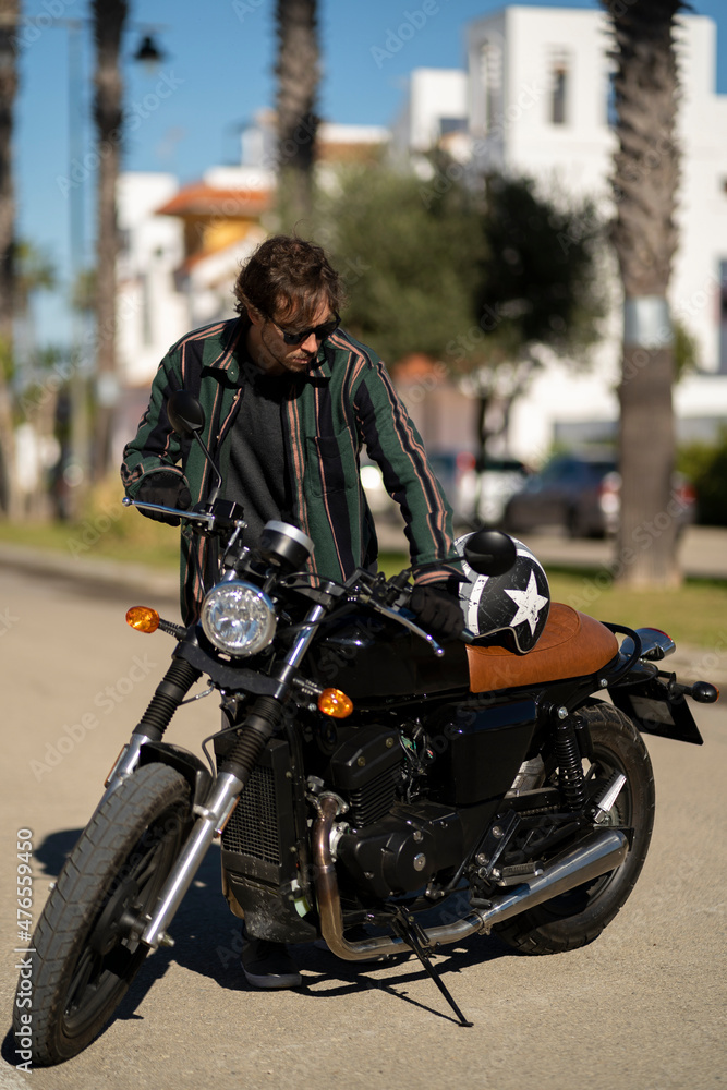 Adolescente hombre joven motero con camisa de cuadros elegante preparándose para viaje. Fondo de palmeras veraniegas. Chico elegante con gafas de sol y moto. Chico poniéndose casco de seguridad.