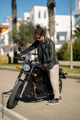 Adolescente hombre joven motero con camisa de cuadros elegante prepar  ndose para viaje. Fondo de palmeras veraniegas. Chico elegante con gafas de sol y moto. Chico poni  ndose casco de seguridad.