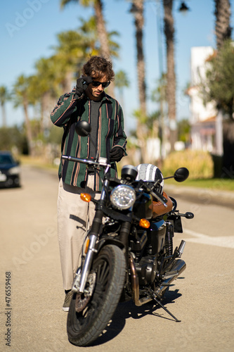 Adolescente hombre joven motero con camisa de cuadros elegante prepar  ndose para viaje. Fondo de palmeras veraniegas. Chico elegante con gafas de sol y moto. Chico poni  ndose casco de seguridad.