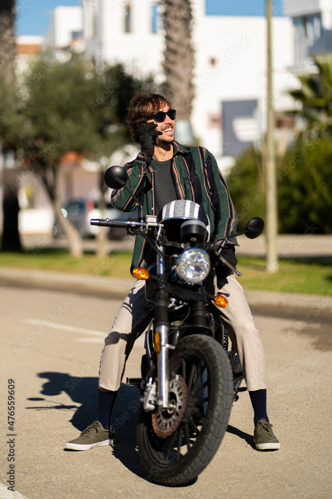 Adolescente hombre joven motero con camisa de cuadros elegante preparándose para viaje. Fondo de palmeras veraniegas. Chico elegante con gafas de sol y moto. Chico poniéndose casco de seguridad.