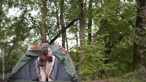 A beautiful woman looks out of the tent and looks around in the woods. Portrait of a tourist on a hike