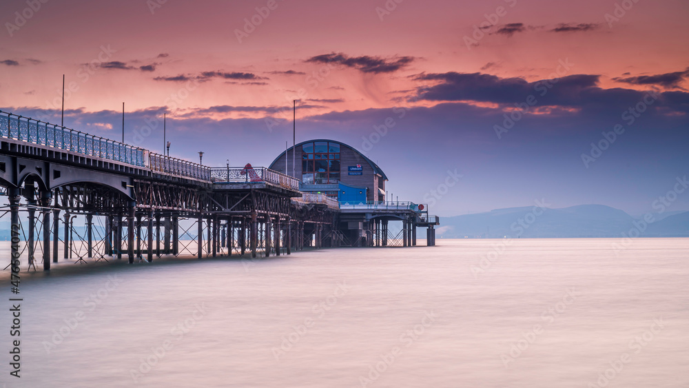 The long pier in the Mumbles, Swansea, south Wales. The wooden and steel structure sits in a calm sea, with a red sky behind it, at sunrise