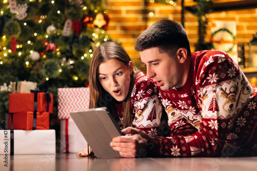 Caucasian couple lying home near Christmas tree background. Beautiful woman with long hair holds digital tablet and emotionally tells funny stories
