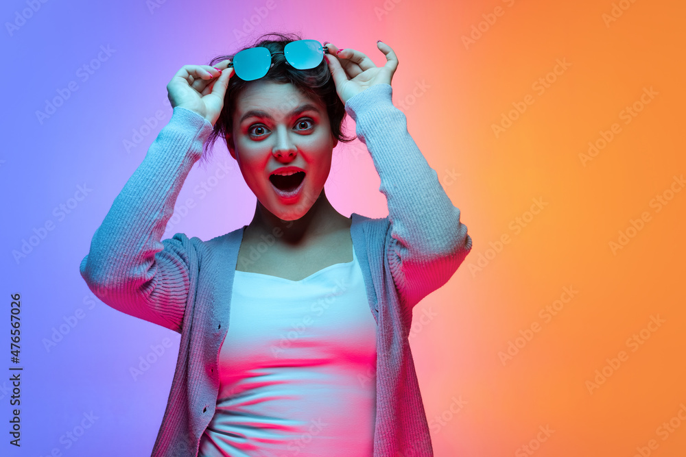 Studio shot of beautiful young girl waering sunglasses isolated on gradient blue-orange color background in neon.