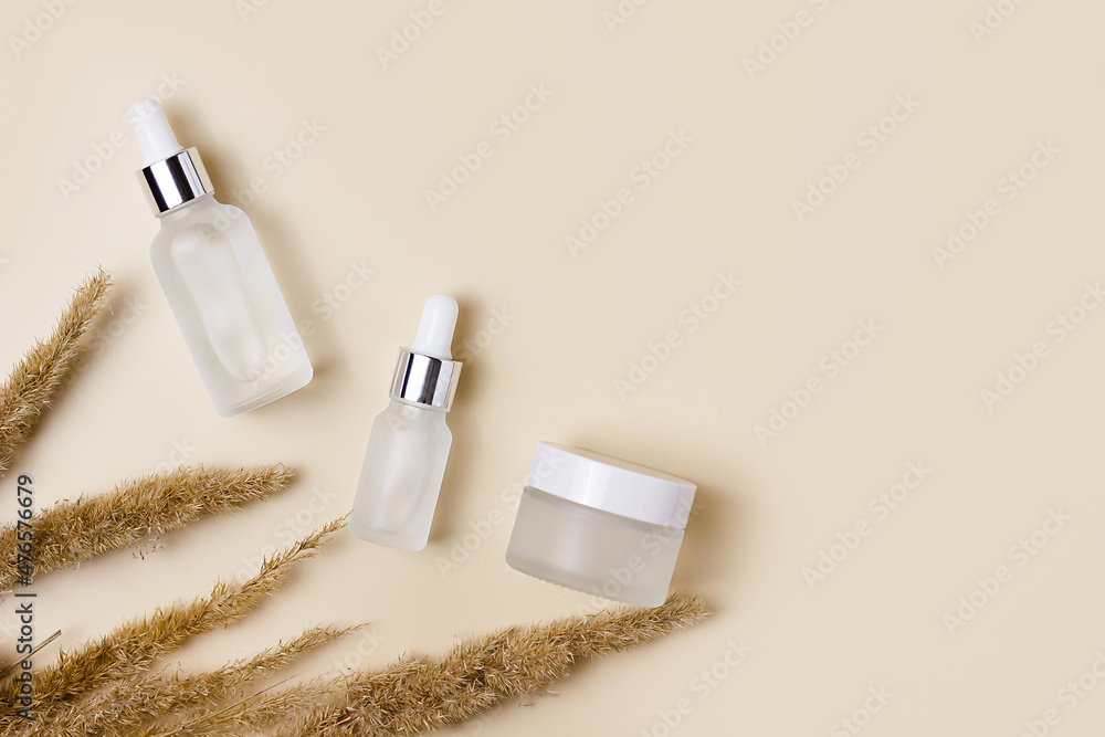 White cosmetic jars on a beige background with dry spikelets of field grass. Winter and autumn care products. Cream, serum, fluid. Minimalism and sustainability. White cosmetic jars on a beige background with dry spikelets of field grass. Winter and autumn care products. Cream, serum, fluid. Minimalism and sustainability.