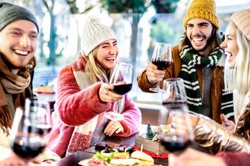 Young friends toasting red wine at restaurant patio - Happy people having fun together at winery bar wearing winter clothes - Dinning life style concept on bright filter with focus on left woman