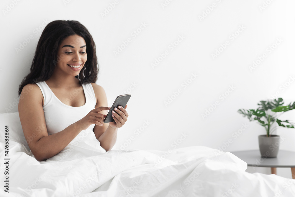 Happy black woman sitting in bed and smiling while reading pleasant message on smartphone in the morning, copy space Happy black woman sitting in bed and smiling while reading pleasant message on smartphone in the morning, copy space