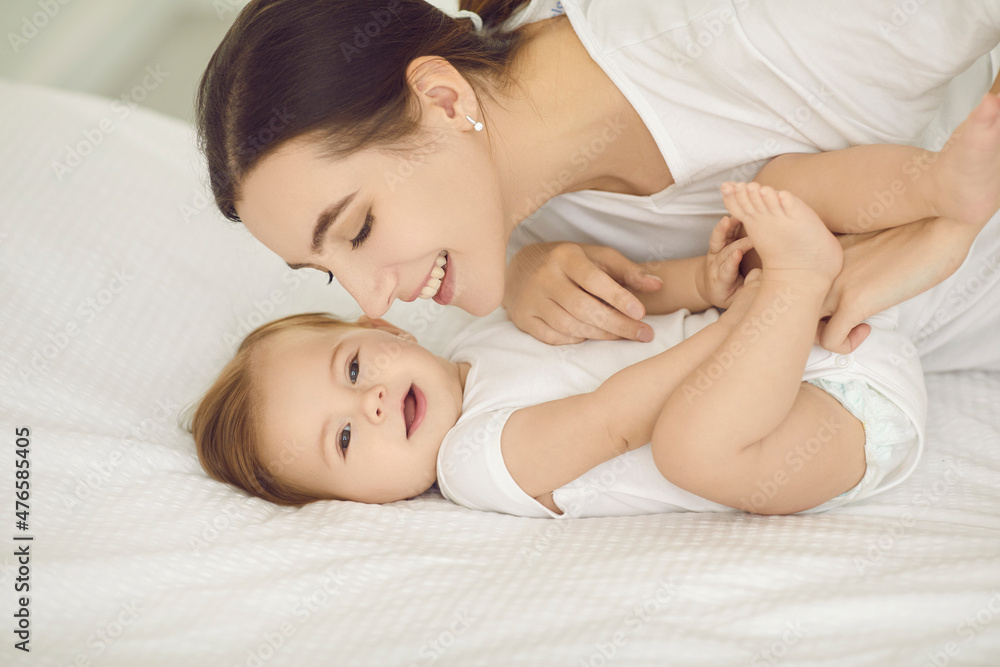 Happy mother and child cuddling on bed. Portrait of smiling young mom playing with cute, beautiful barefoot baby wearing fresh white diaper in nursery room or bedroom. Family, love, and care concept Happy mother and child cuddling on bed. Portrait of smiling young mom playing with cute, beautiful barefoot baby wearing fresh white diaper in nursery room or bedroom. Family, love, and care concept