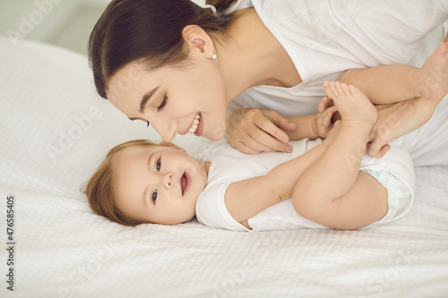 Happy mother and child cuddling on bed. Portrait of smiling young mom playing with cute, beautiful barefoot baby wearing fresh white diaper in nursery room or bedroom. Family, love, and care concept