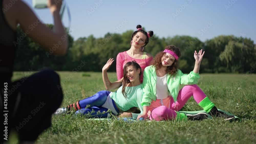 Wide shot three retro women posing on green summer sunny lawn as friend taking photo on camera. Happy confident slim beautiful Caucasian friends outdoors smiling sitting on meadow Wide shot three retro women posing on green summer sunny lawn as friend taking photo on camera. Happy confident slim beautiful Caucasian friends outdoors smiling sitting on meadow