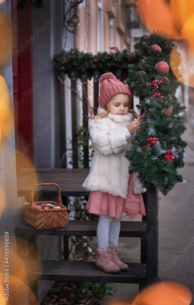  Little girl in a white fur coat dresses up the porch and the front door for the new year and christmas. Preparing for the new year. home decoration for the holiday. Christmas background. 
