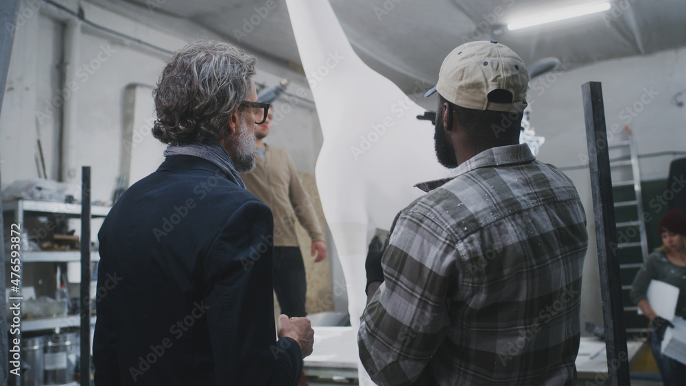 Low angle of mature customer in suit discussing gypsum giraffe statue with black craftsman in illuminated workroom Low angle of mature customer in suit discussing gypsum giraffe statue with black craftsman in illuminated workroom