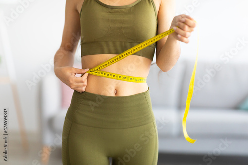Cropped view of slim Indian woman measuring her waist with tape measure at home, closeup photo