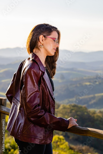 Young woman on a mountain gazebo looking at the landscape with hills and native vegetation and clouds in the sky on the horizon. With the golden light of Sunset or Sunrise on your back and hair.