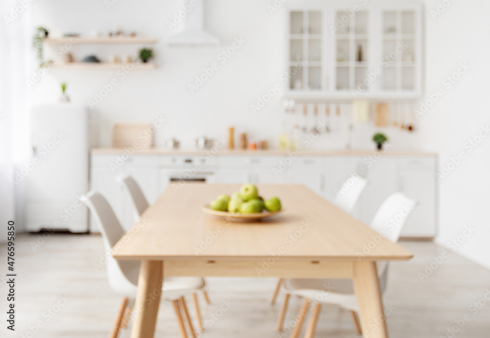Blurred background with modern light kitchen, wooden dining table and white furniture, chairs, kitchenware and utensils