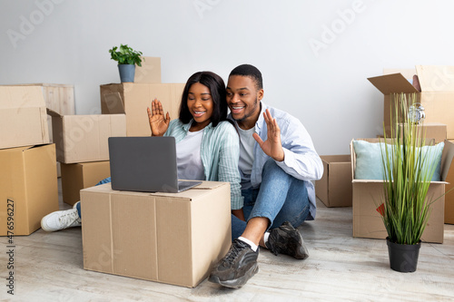 Young black family chatting online via laptop, sitting among carton boxes in new apartment and waving hands to camera