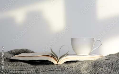 The pages in the book lying on the sweater are folded in the shape of a heart. Next to a cup of coffee. White background. Concept - morning coffee