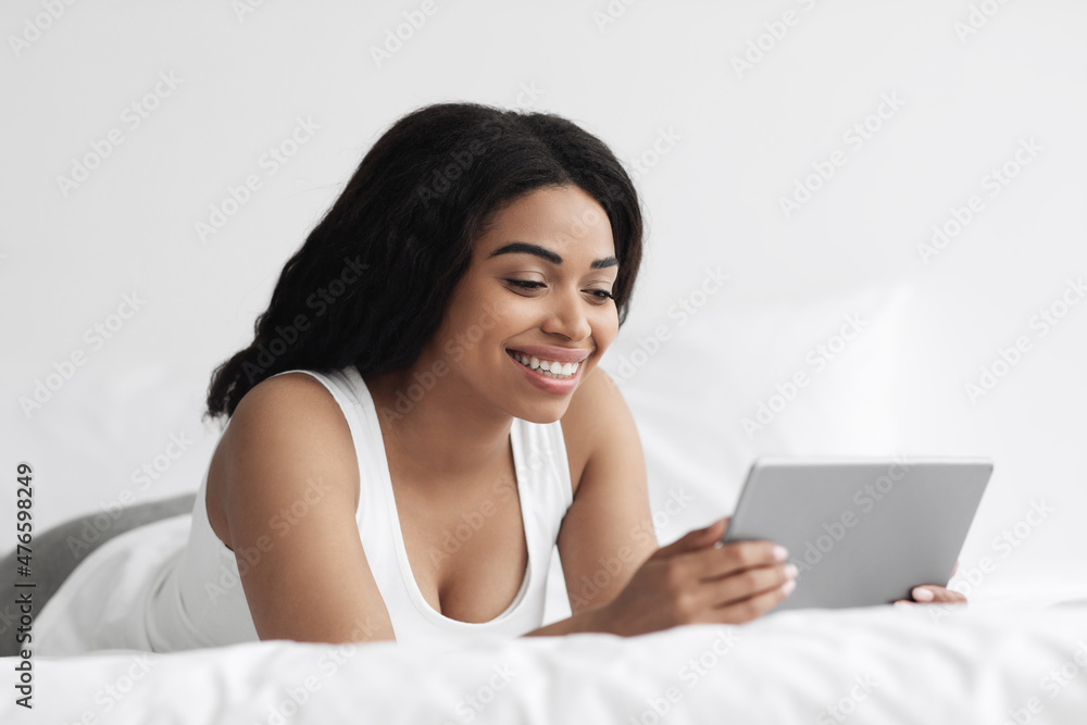Watching video and news. Excited black woman holding digital tablet and lying on bed in the morning, free space Watching video and news. Excited black woman holding digital tablet and lying on bed in the morning, free space