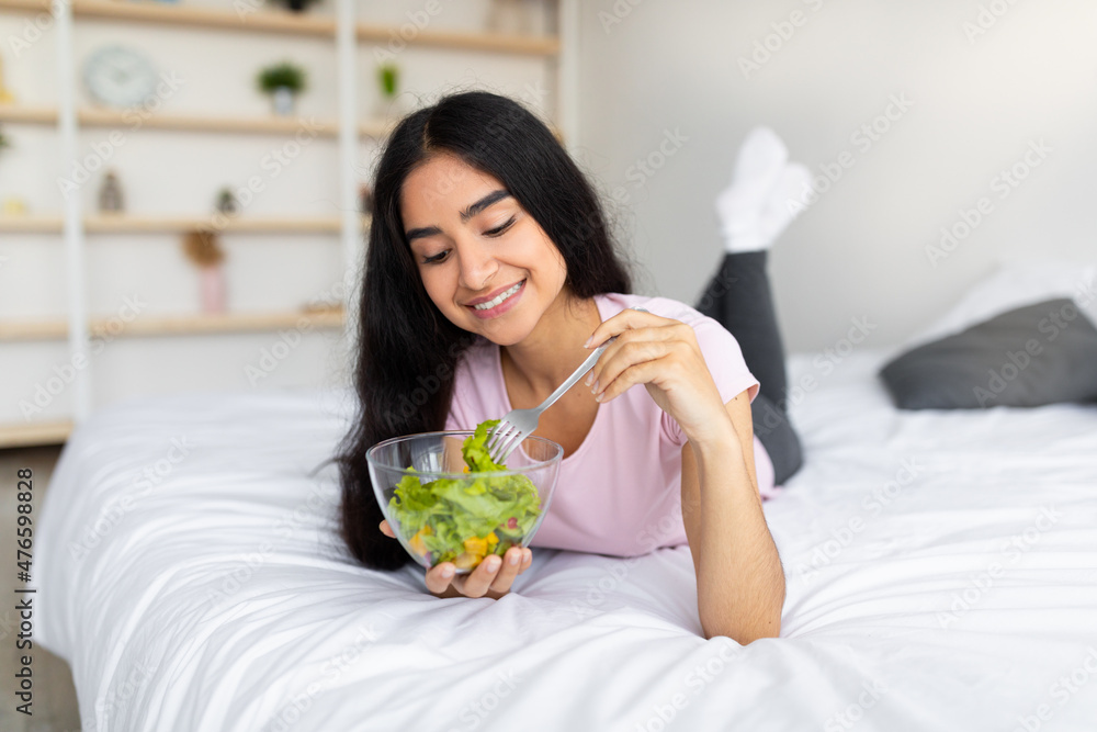 Slimming diet concept. Lovely Indian lady eating tasty vegetable salad on comfy bed at home Slimming diet concept. Lovely Indian lady eating tasty vegetable salad on comfy bed at home