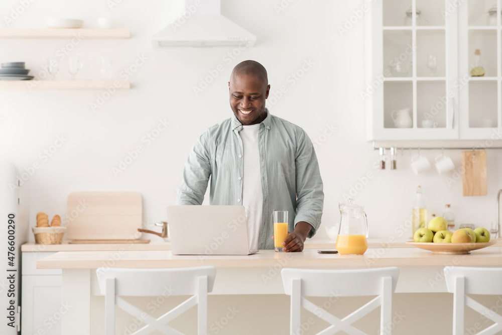 Positive black mature man reading online news on laptop and drinking orange juice in kitchen, enjoying morning at home Positive black mature man reading online news on laptop and drinking orange juice in kitchen, enjoying morning at home