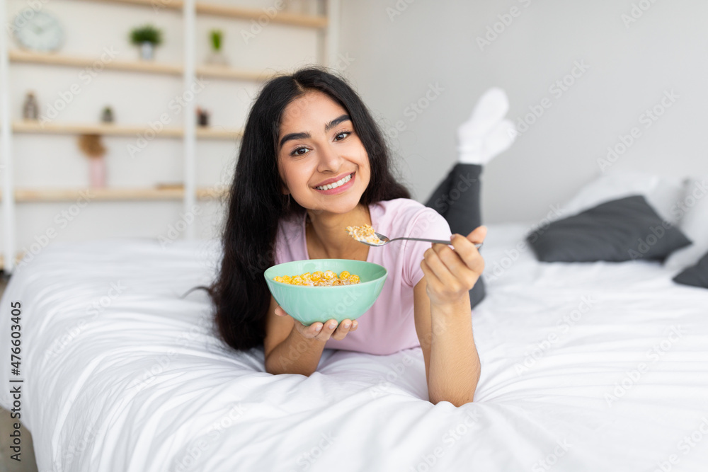 Balanced diet for weight loss. Full length of lovely Indian lady eating cereal with milk while lying on bed at home Balanced diet for weight loss. Full length of lovely Indian lady eating cereal with milk while lying on bed at home