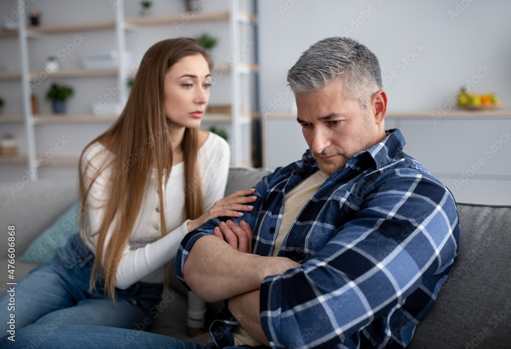 After family conflict. Upset mature man sitting on couch, his wife comforting him, asking for forgiveness at home After family conflict. Upset mature man sitting on couch, his wife comforting him, asking for forgiveness at home