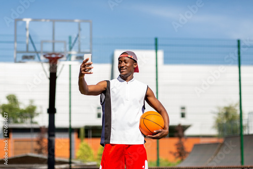 Portrait of black basketball player holding ball, taking selfie on smartphone at outdoor sports arena © Prostock-studio