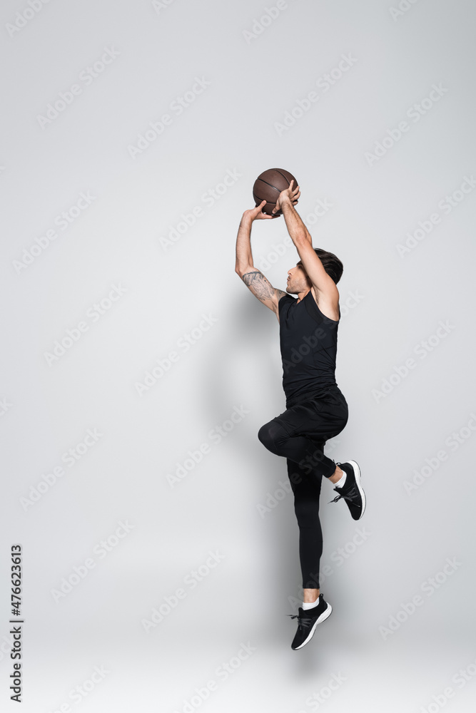 Obraz premium Side view of tattooed sportsman with basketball ball jumping on grey background