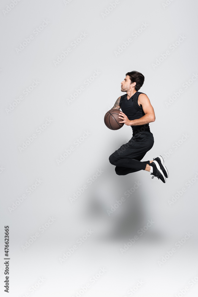 Fototapeta premium Side view of tattooed sportsman with basketball ball jumping on grey background