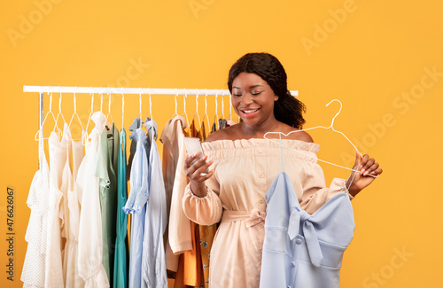 Happy black woman using smartphone for online shopping, choosing dress near clothing rail, picking summer wardrobe