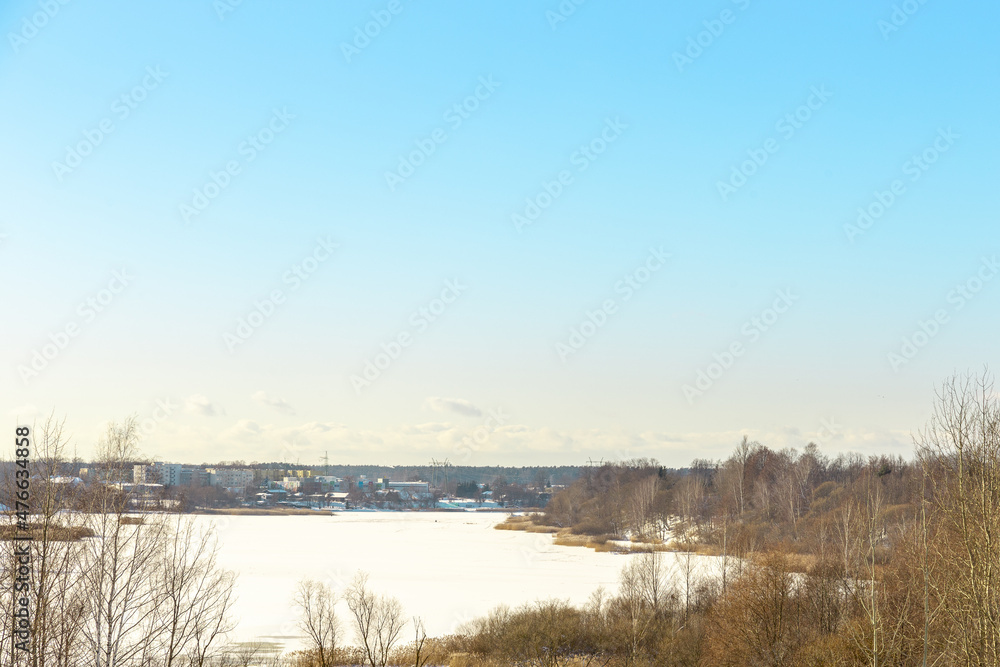 Panoramic winter landscape view with frozen river or lake covered with ice, reeds and some trees off the coast, small village and tower in background