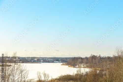 Panoramic winter landscape view with frozen river or lake covered with ice, reeds and some trees off the coast, small village and tower in background © Photo by ERIKS ROZE