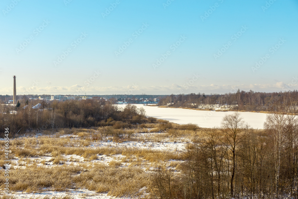 Panoramic winter landscape view with frozen river or lake covered with ice, reeds and some trees off the coast, small village and tower in background Panoramic winter landscape view with frozen river or lake covered with ice, reeds and some trees off the coast, small village and tower in background