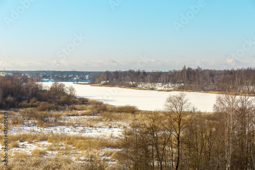 Panoramic winter landscape view with frozen river or lake covered with ice, reeds and some trees off the coast, small village and tower in background © Photo by ERIKS ROZE