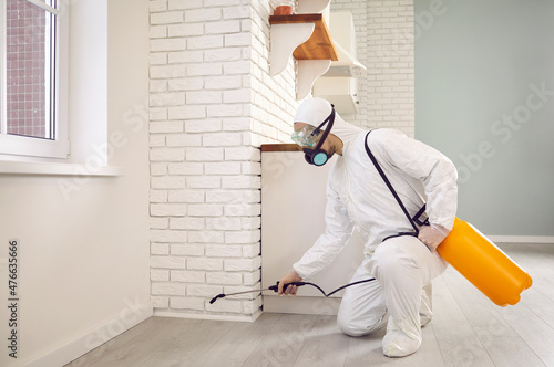 Pest control guy inside the house. Male in white protective overalls crouching by kitchen wall and spraying cockroach insecticide for maintaining clean, safe and insect free living environment at home photo