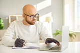 Serious man in glasses and comfortable fingerless compression hand gloves for easing arthritis pain sitting at desk, using computer, typing on keyboard, holding pen and taking study notes on paper