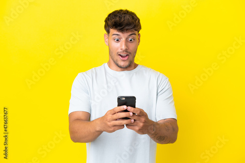 Young caucasian handsome man isolated on yellow background looking at the camera while using the mobile with surprised expression © luismolinero