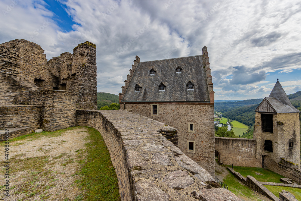 Upper courtyard in the outdoor ruins of the medieval castle of Bourscheid, ruined walls, a building and the watchtower, hills covered with lush green trees in the background, Luxembourg