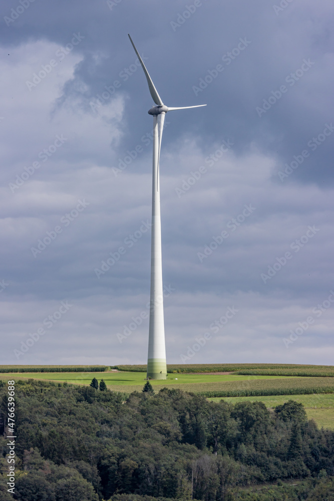 Huge windmill on farm land surrounded by lush green trees with a blue sky in the background, white metal pole and vanes, sunny summer day in Luxembourg Huge windmill on farm land surrounded by lush green trees with a blue sky in the background, white metal pole and vanes, sunny summer day in Luxembourg