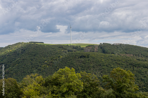Countryside with a hill covered with lush green trees with a windmill with a cloud covered sky in the background, white metal pole and blades, sunny summer day in Luxembourg