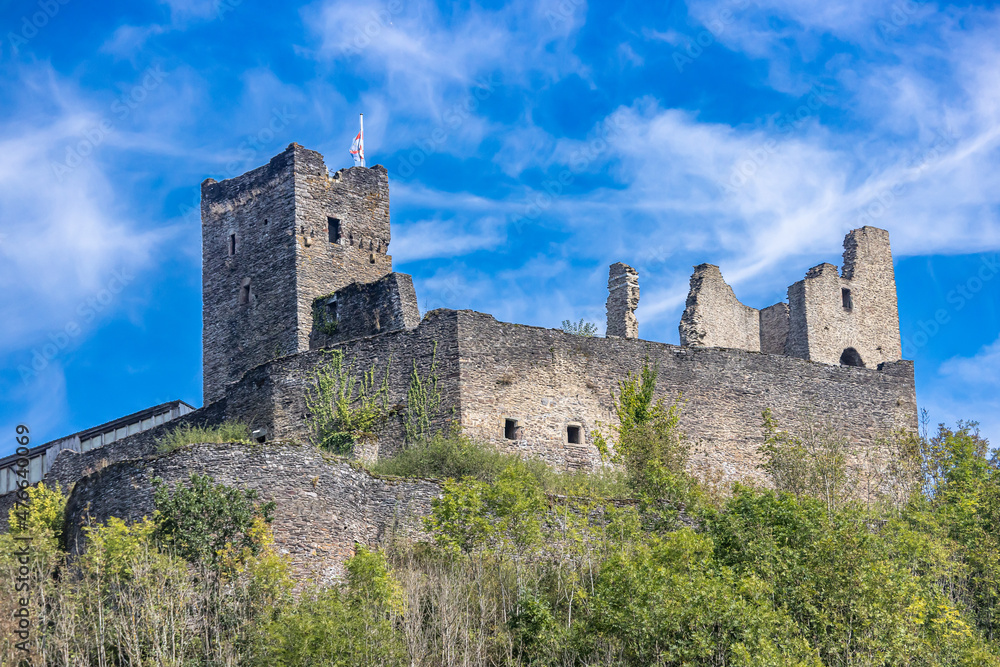 Ruined Brandenburg Castle on a promontory surrounded by small green trees, towers and stone walls, blue sky and few white clouds in the background, sunny summer day in Luxembourg