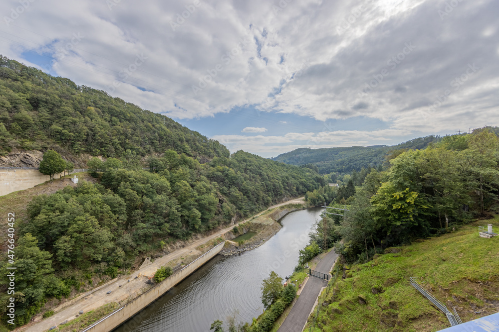 River Esch-sur-Sure, a closed country road between hills with lush green trees and wild vegetation with the valley in the background, cloudy day with a sky with abundant clouds in Luxembourg