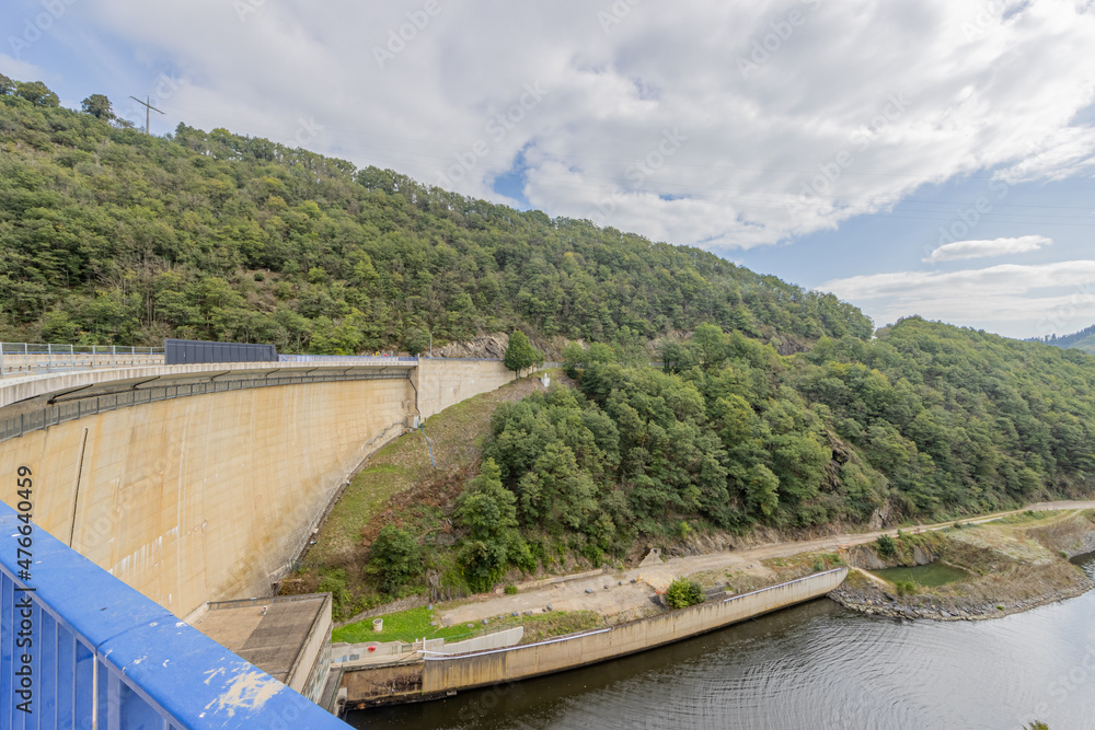 Wall of the Esch-sur-Sure dam with hills with lush green trees and wild vegetation in the background, cloudy day with a sky with abundant clouds in Luxembourg