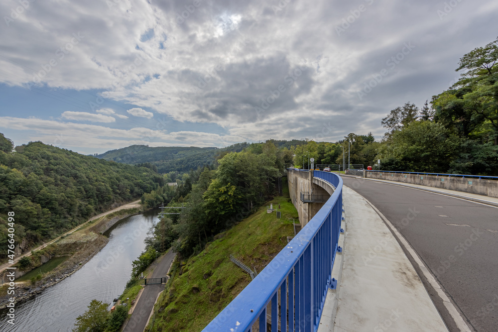 Obraz premium Rural road closed for traffic next to the river Esch-sur-Sure, hills covered with lush green trees and wild vegetation in the background, blue metal fence, cloudy day with clouds in Luxembourg