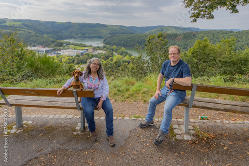 Smiling tourist couple with their dog, wooden bench on a viewpoint, the Esch-sur-Sure lake and the dam surrounded by wild vegetation and lush green trees, looking at camera, sunny day in Luxembourg
