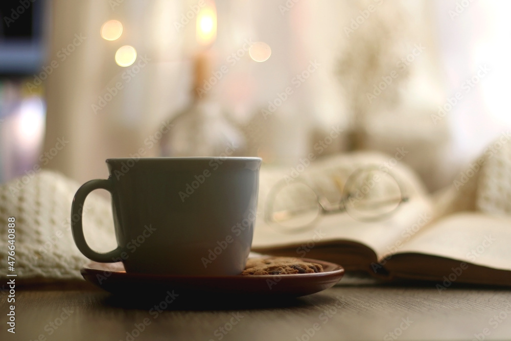 Cup of warm drink, chocolate chip cookies, open book and reading glasses. Candles, flowers and bokeh lights in the background. Hygge at home. Selective focus.